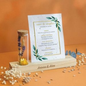 Support floral avec tube en verre contenant des fleurs séchées et plateau en bois soutenant une carte en papier ensemencé servant de support de communication interne entreprise.